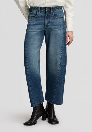 Personne portant un jean bleu taille haute à jambes larges avec des ourlets bruts et des bottines en cuir noires, debout devant un fond uni.