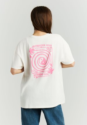 Vrouw met bruin haar, die een wit T-shirt draagt met een roze spiraal en de tekst "Saltwater Soul Balanced Roll" op de achterkant, gecombineerd met een blauwe spijkerbroek.