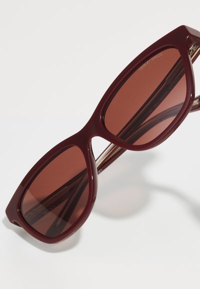 Ochelari de soare burgundy cu lentile maronii, formă unghiulară și rame din plastic lucios, prezentând accente metalice și branding pe braț.