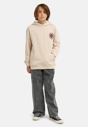 Beżowa bluza z kapturem z przednią kieszenią i pomarańczowym detalem graficznym. Połączona z luźnymi szarymi dżinsami i czarnymi trampkami z białymi podeszwami.