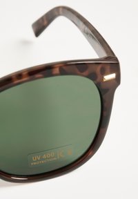 Occhiali da sole tartaruga marrone con lenti verdi, dotati di etichetta di protezione UV 400. Texture liscia con un design a forma rettangolare.
