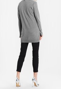 Maglione grigio a maniche lunghe, pantaloni neri slim con fessure alla caviglia e tacchi alti argentati. Texture liscia, design casual, minimalismo nei dettagli.