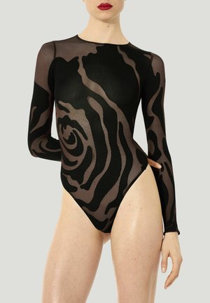 Donna che indossa un body nero trasparente a maniche lunghe con motivi astratti vorticosi, in piedi con una mano dietro la schiena.