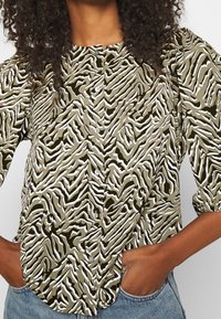 Camisa verde oliva con rayas de cebra texturizadas en blanco. Presenta botones en la parte delantera y mangas abullonadas, con un corte relajado.