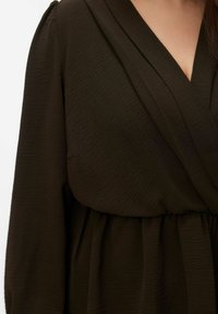 Donkerbruine jurk met een gestructureerde, geplooide stof. Heeft een wikkelontwerp en een gerimpelde taille. Lange mouwen met een lichte pof bij de schouders.