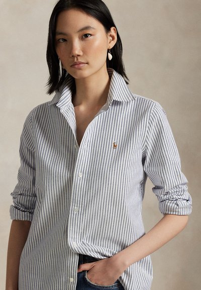 Kobieta o prostych czarnych włosach nosi niebiesko-białą koszulę w paski z guzikami, podwiniętymi rękawami oraz małym haftowanym logo.