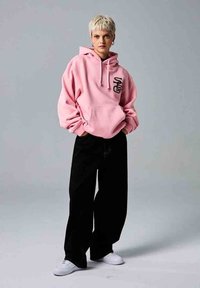 Sudadera rosa con un bolsillo frontal y texto gráfico negro, combinada con pantalones holgados negros y zapatillas blancas sobre un fondo liso.
