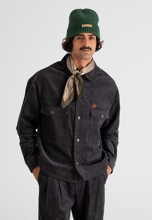 Man with mustache wearing green knit beanie, beige patterned neckerchief, dark corduroy jacket, and matching pants, standing with hands in pockets.