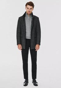 Szary wełniany płaszcz z klapami, dwoma przednimi kieszeniami, szary sweter pod spodem, czarne dopasowane spodnie i czarne buty. Neutralne tło.