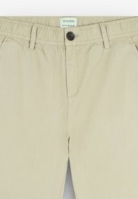 Pantalones de mezcla de algodón color beige con una cintura elástica, cierre frontal de botón y bolsillos laterales. Textura suave y diseño ajustado.