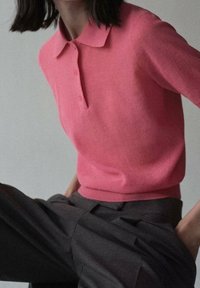 Jasnoróżowa koszulka polo wykonana z miękkiej tkaniny, z kołnierzykiem i trzema guzikami, zestawiona z ciemnoszarymi plisowanymi spodniami. Minimalistyczny design.