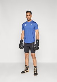 Μπλε αθλητική μπλούζα με υφασμάτινο σχέδιο, μαύρο σορτς, μαύρα γάντια πυγμαχίας και μαύρα με καφέ ψηλά αθλητικά παπούτσια. Στάση όρθια.