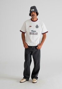 Vīrietis ar ūsām, ģērbts pelēkā beanie cepurē, baltā T-kreklā ar tekstu "Escape Berlin", brīvos melnajos džinsos un bēšās sporta apavās, stāvot pret vienkāršu fona.