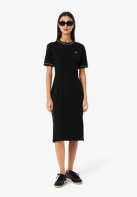 Musta mekko, joka on valmistettu jersey-kankaasta, jossa on lyhyet hihat. Mekossa on pyöreä kaula-aukko, jossa on vihreä yksityiskohta, ja se on slim-fit -mallinen. Yhdistetty mustiin lenkkareihin.