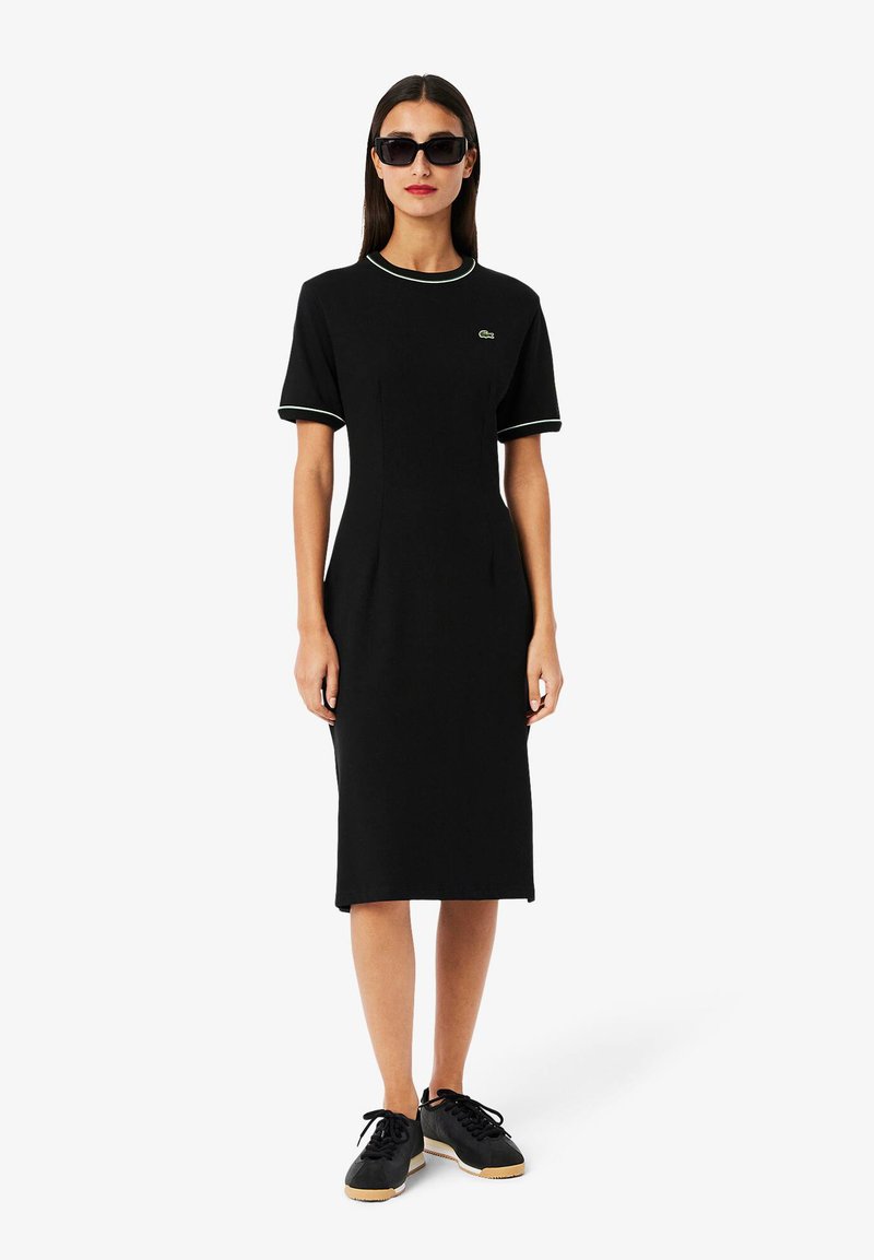 Musta mekko, joka on valmistettu jersey-kankaasta, jossa on lyhyet hihat. Mekossa on pyöreä kaula-aukko, jossa on vihreä yksityiskohta, ja se on slim-fit -mallinen. Yhdistetty mustiin lenkkareihin.