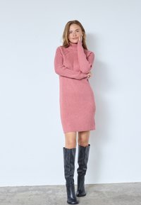 Rosa ribbad stickad klänning med hög hals, slim fit och långa ärmar, tillsammans med knähöga svarta läderstövlar.
