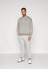 Γκρι φούτερ με στρογγυλή λαιμόκοψη και διακριτικό λογότυπο, συνδυασμένο με ανοιχτό γκρι φόρμας. Λευκά αθλητικά παπούτσια με μαύρες λεπτομέρειες. Απλός, μοντέρνος σχεδιασμός.