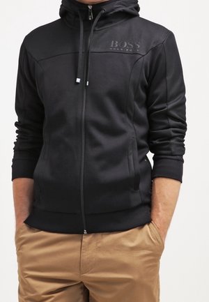 Mann trägt einen schwarzen Kapuzenpullover mit Reißverschluss und dem Logo „BOSS HUGO BOSS“ auf der Brust sowie beige Hose, Hände in den Taschen, steht vor weißem Hintergrund.