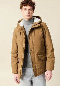 Joven vestido con una chaqueta de invierno con capucha color beis sobre un suéter de cuello alto gris y jeans azul claro, de pie contra un fondo beige.