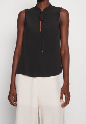 Blouse noire sans manches avec détail à nouer au col, associée à un pantalon ample de couleur claire sur une personne à la peau foncée.