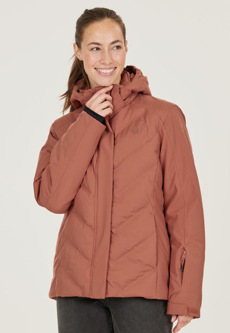 Veste rembourrée de couleur rouille avec une capuche, présentant des coutures diagonales, des poignets noirs en accent et une poche zippée sur la manche gauche.