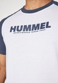Bílá tričko s námořnickými raglánovými rukávy, s výrazným logem "HUMMEL" v tmavě modré barvě a textem "SPORTSWEAR SINCE 1923" pod ním. Bavlněný materiál.