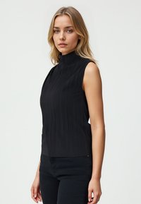 Zwarte mouwloze top met een hoge opstaande kraag, voorzien van een geweven ribpatroon en een getailleerde pasvorm. Gecombineerd met zwarte broek.