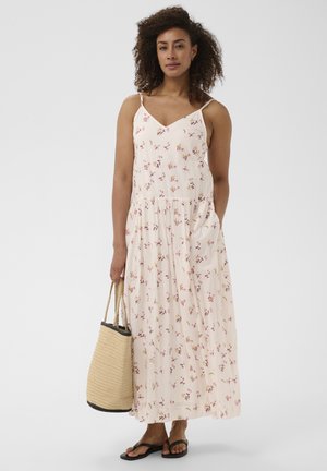 Femme aux cheveux bouclés portant une robe longue fleurie sans manches, des sandales noires et tenant un sac fourre-tout en paille tressée.