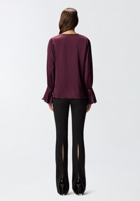 Burgundijska svilenka s dugim raskošnim rukavima i detaljima na gumbima, u kombinaciji s crnim prilagođenim hlačama s bočnim prorezima i štiklama.