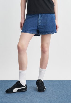 Személy kék farmer rövidnadrágban, fehér bokazokniban, fekete-fehér Puma tornacipőben és fekete pólóban, kék szőnyegen állva.