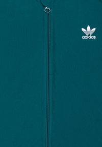 Tirkīza krāsas zip-up jaka, kas izgatavota no viegla auduma, ar centrālo rāvējslēdzēju un izšuvumu balto Adidas logo uz krūškurvja.