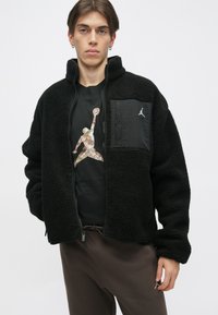 Fekete bolyhos kabát magas gallérral, cipzárral, és fekete zsebbel, amelyen logó található. Fekete póló és barna nadrág fölött viselve.