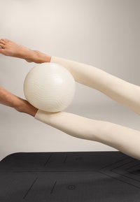 Bela fitnes žoga, uravnotežena med dvema nogama, oblečenima v tesne, teksturirane kremne legice. Črna vadbena podloga z subtilnimi vzorci spodaj.