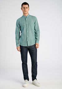 Lichtgroen overhemd met lange mouwen, subtiele textuur; gecombineerd met donkere spijkerbroek en witte sneakers, staand voor een neutrale achtergrond.