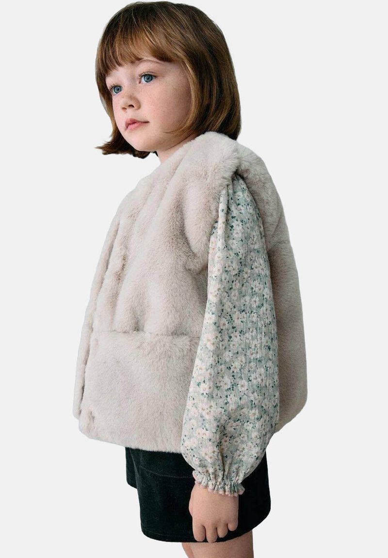 Lažni krzneni prsluk u svijetloj bež boji, s pahuljastom teksturom. Uparen s bluzom dugih rukava s cvjetnim uzorkom i crnim šortsevima.