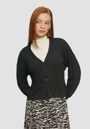 Cardigan noir texturé avec un design en V et trois grands boutons. Porté sur un col roulé léger et une jupe à motifs.