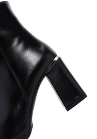 Bottine en cuir noir avec une finition brillante, dotée d'un talon carré avec un accent métallique et d'une texture lisse.