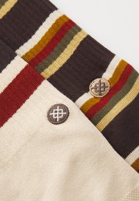 Strikket tekstil med horisontale striper i rød, gul, grønn og kremfarge, med en strukturert brun seksjon og to broderte knapper.