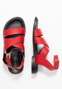 Punaiset nahkasaappaat, joissa on ristikkäiset remmit, musta pohja ja kultaiset niitit pohjan ympärillä. Mukana kultainen solki säätöä varten.