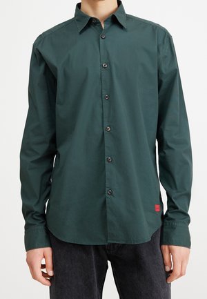 Person trägt ein dunkelgrünes, langärmliges Hemd mit Knöpfen und schwarze Jeans, steht vor einem schlichten weißen Hintergrund.