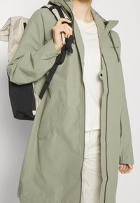 Veste imperméable vert clair avec capuche, fermeture éclair à l'avant et poches. Portée par-dessus un haut crème, associée à un sac à dos noir et beige.