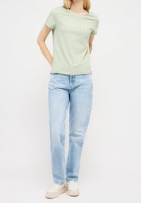 Camiseta de algodón verde claro con estampado "MUSTANG", combinada con jeans de mezclilla azul claro y zapatillas blancas. Mangas cortas y corte relajado.