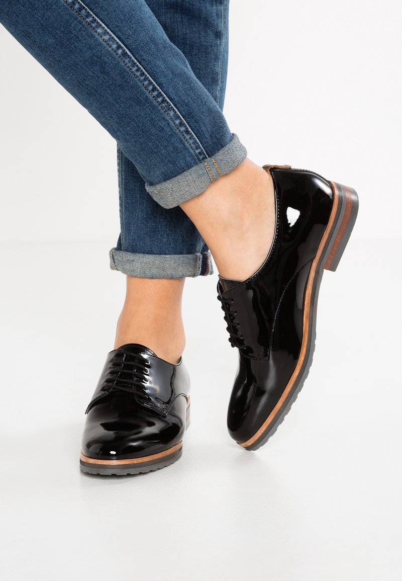 Chaussures en cuir verni noir avec un bout rond, des lacets, un talon en bois et une semelle en caoutchouc, portées avec un jean denim retroussé.