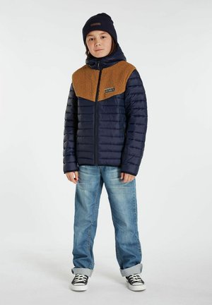 Donkerblauwe pufferjack met een bruine fleece bovenkant, voorrits en patchdetail. Gecombineerd met lichtblauwe jeans en zwarte sneakers.