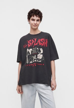 Personne portant un t-shirt graphique noir ample avec un motif tropical rouge et blanc et du texte, associé à un pantalon gris clair, sur un fond uni.