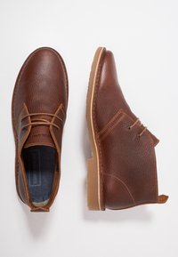 Hnědé kožené boty s texturovaným povrchem, s šněrováním, kulatou špičkou a světlými gumovými podrážkami. Jsou viditelné akcentové prošívání.