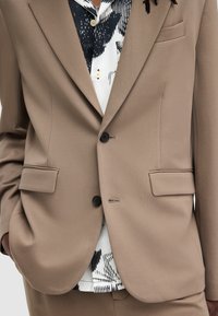 Blazer marrón claro con dos bolsillos frontales y botones negros, superpuesto a una camisa de patrón blanco y negro. Textura suave, corte ajustado.