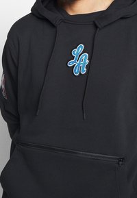 Sudadera negra con un logo bordado en azul de "LA", cordones ajustables y un bolsillo frontal con cremallera. Hecha de un tejido suave con una textura lisa.