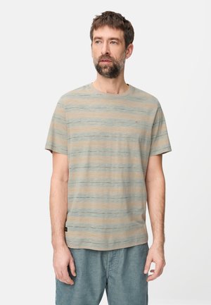 Mann mit Bart, der ein hellgraues und beige gestreiftes T-Shirt mit kurzen Ärmeln und blaue Hosen trägt, steht vor einem schlichten weißen Hintergrund.