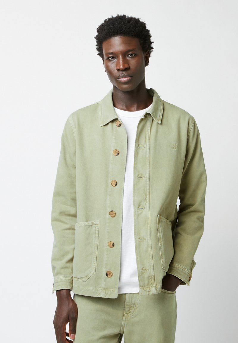 Lichtgroene denim jacket met een kraag, houten knopen, twee frontzakken en een relaxte pasvorm, gedragen over een wit T-shirt.